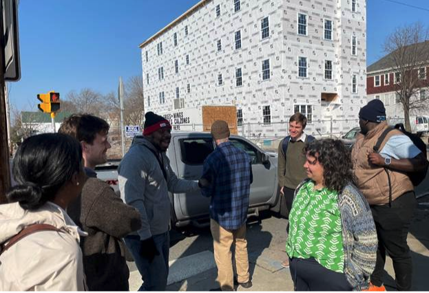 seven people talk outside as a pickup truck drives by in the background